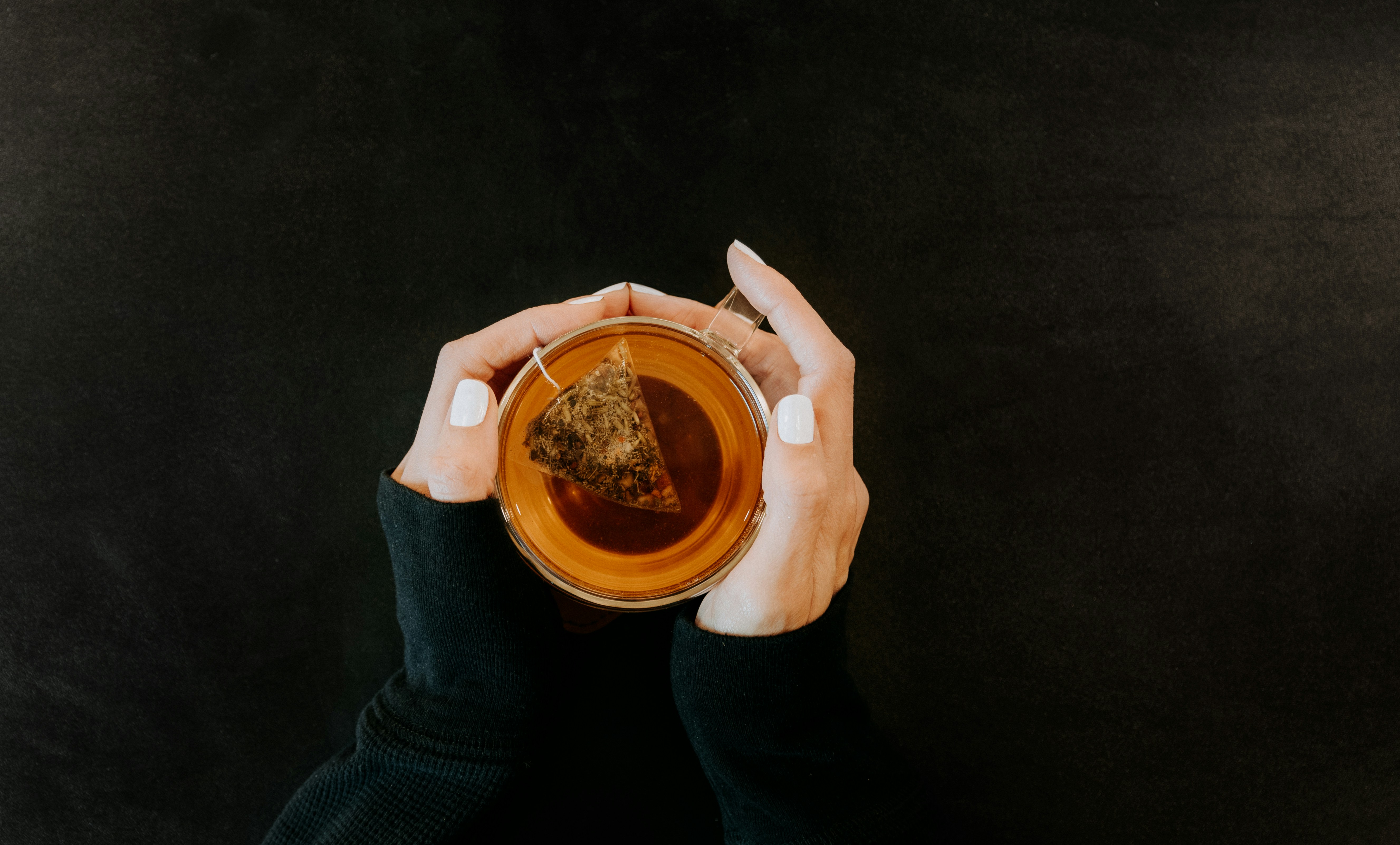 Herbal tea bag steeping in a glass cup held over a dark table