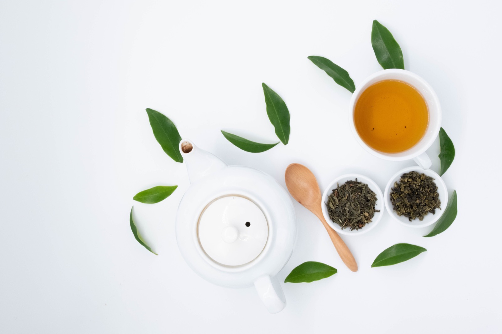 Green tea cup, teapot, and bowls of loose green tea leaves on a white surface